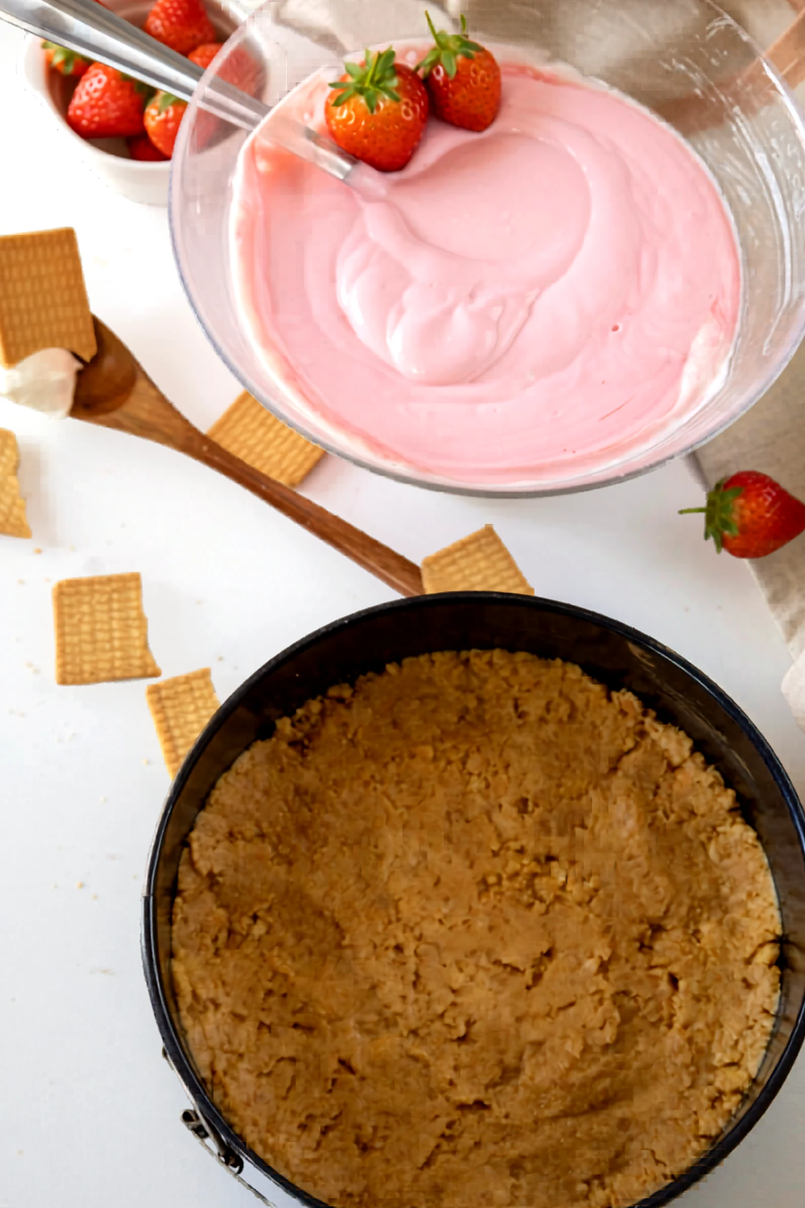 Pink strawberry cheesecake filling in a mixing bowl beside the crust.